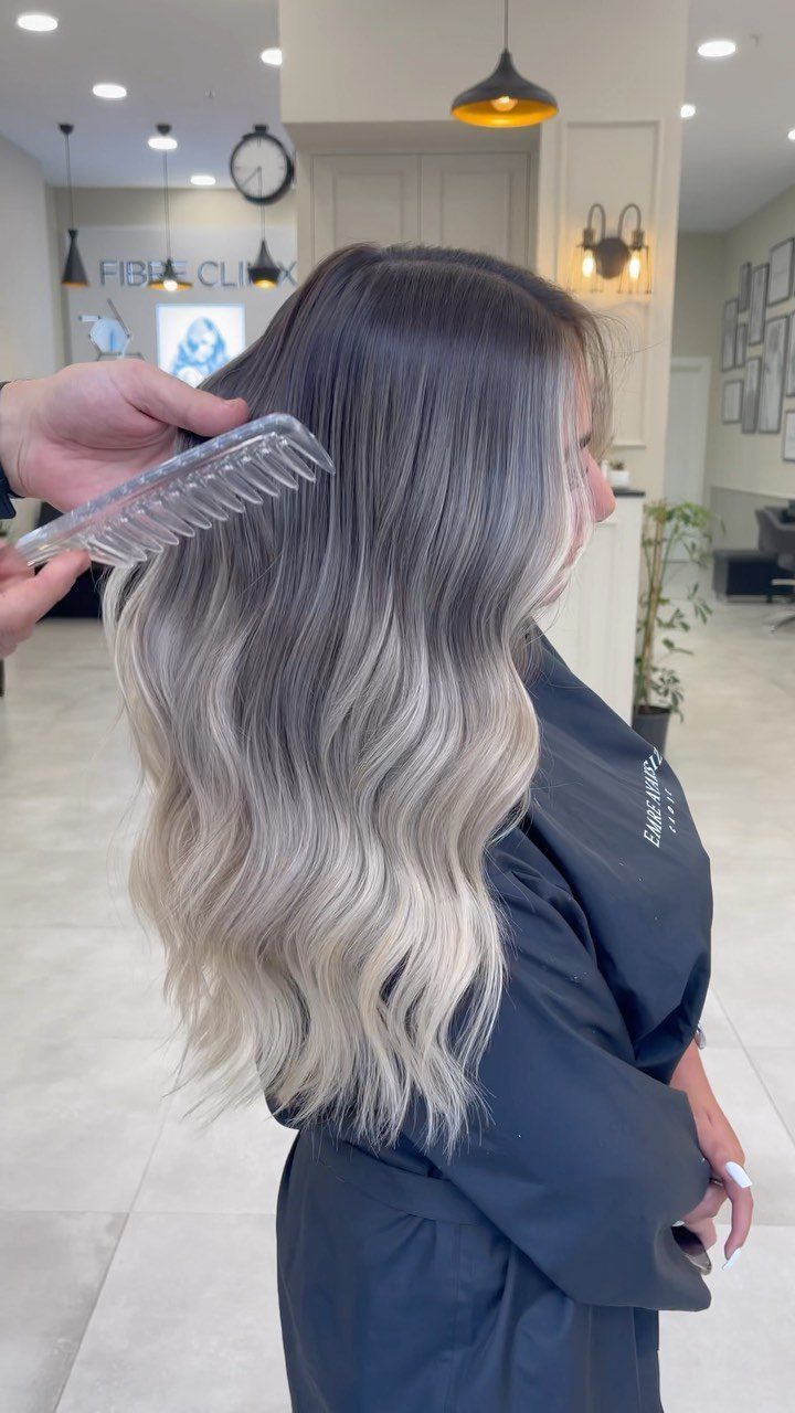 Wavy ash-blonde hair fades from dark roots to pale ends in a salon chair