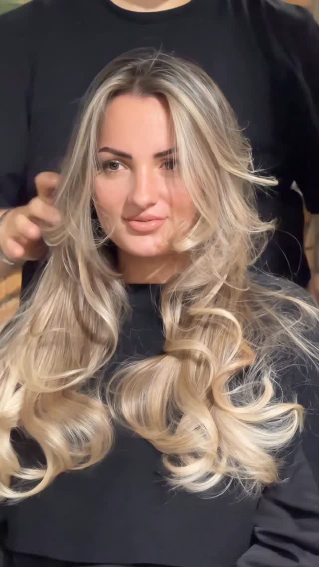 Woman displaying long ash blonde balayage with dark roots and curled ends