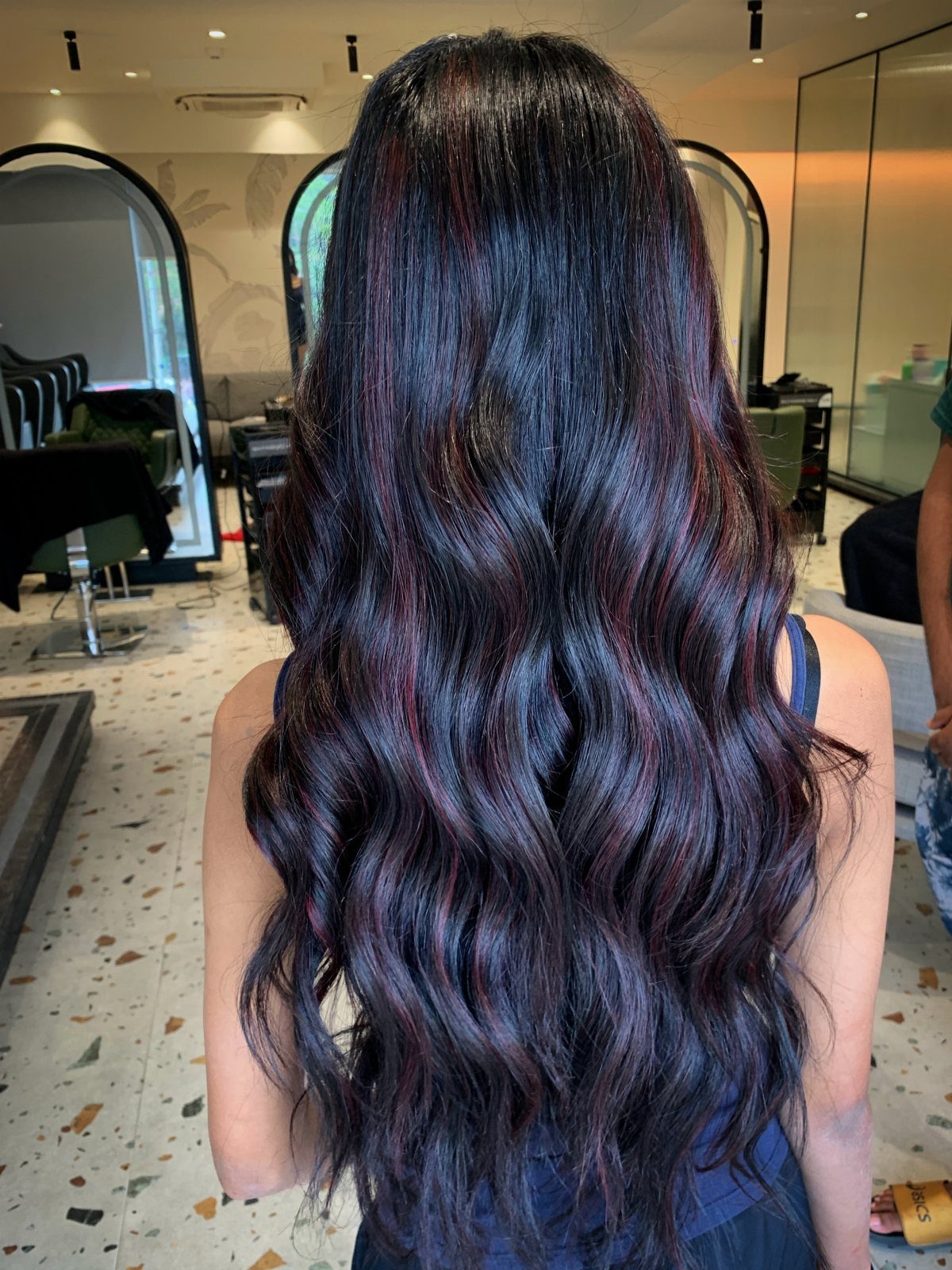 Long black waves show subtle burgundy-red balayage highlights in a salon