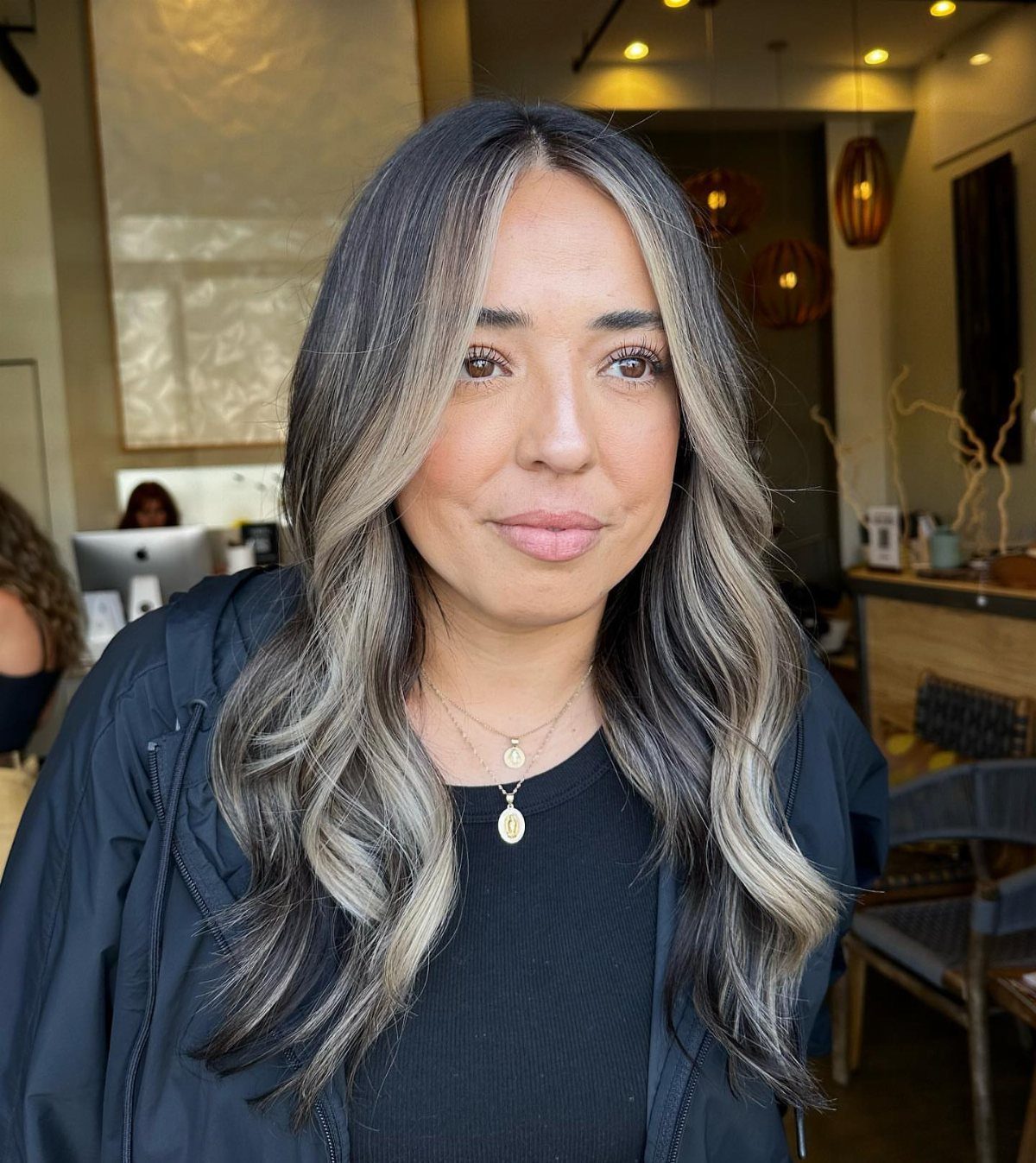 Woman with long wavy black hair and light brown highlights poses indoors
