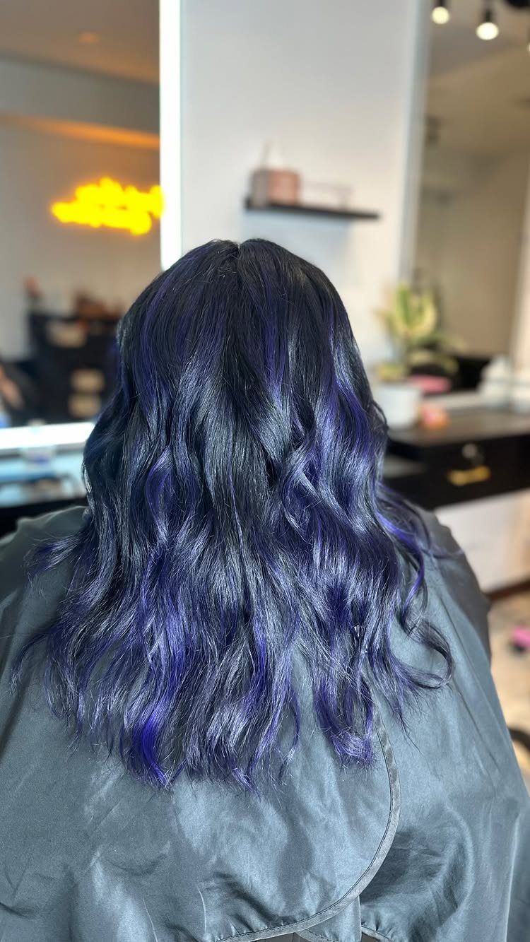 Long dark waves with vivid blue-purple highlights over a gray salon cape