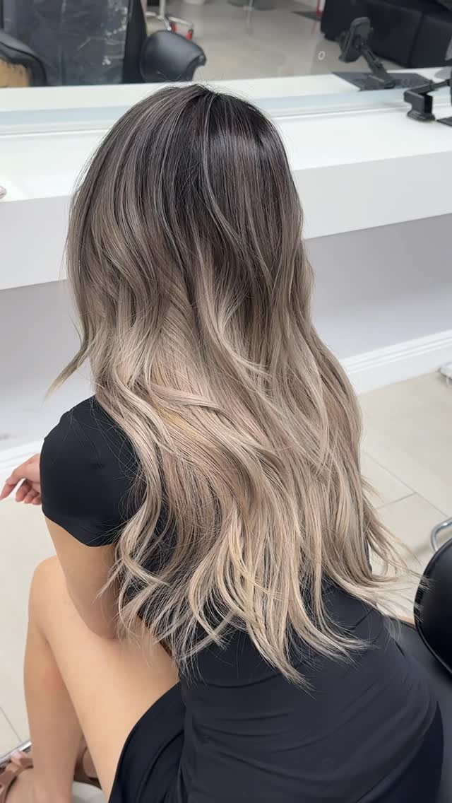 Long wavy hair blends cool dark roots into pale beige-blonde ribbons in a salon