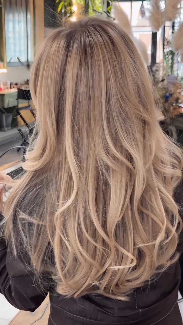 Long blonde balayage waves with darker roots, photographed from behind in a salon.