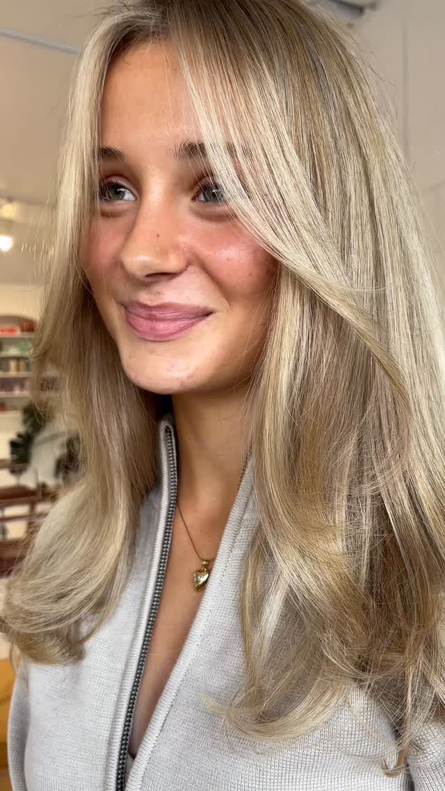 Woman with long, warm blonde hair and darker roots wears a light gray top