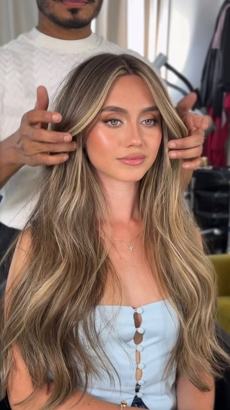 Woman displays long blonde hair with darker roots and golden highlights in a salon.