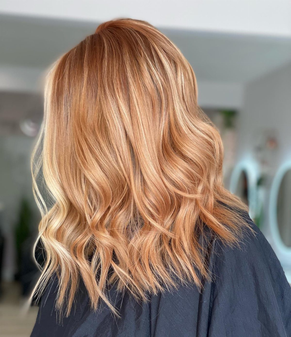 Long wavy strawberry blonde hair with bright balayage highlights over a salon cape