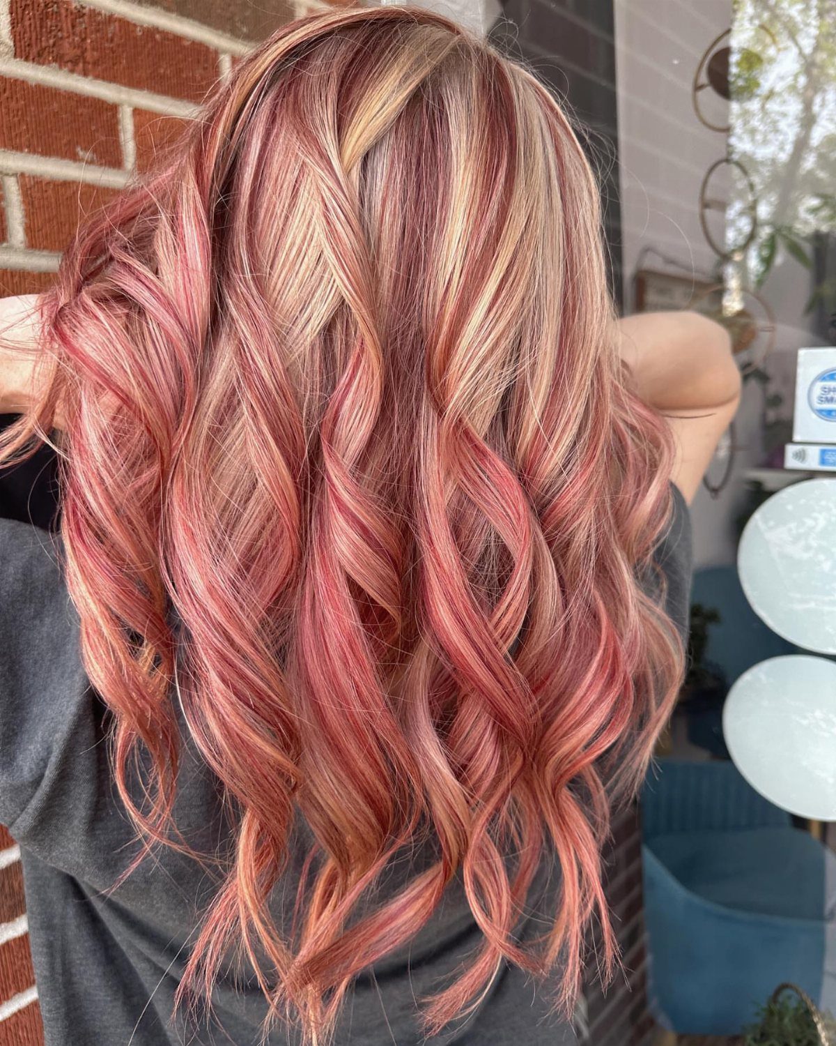Long rose-red curls with pale blonde highlights against a brick wall