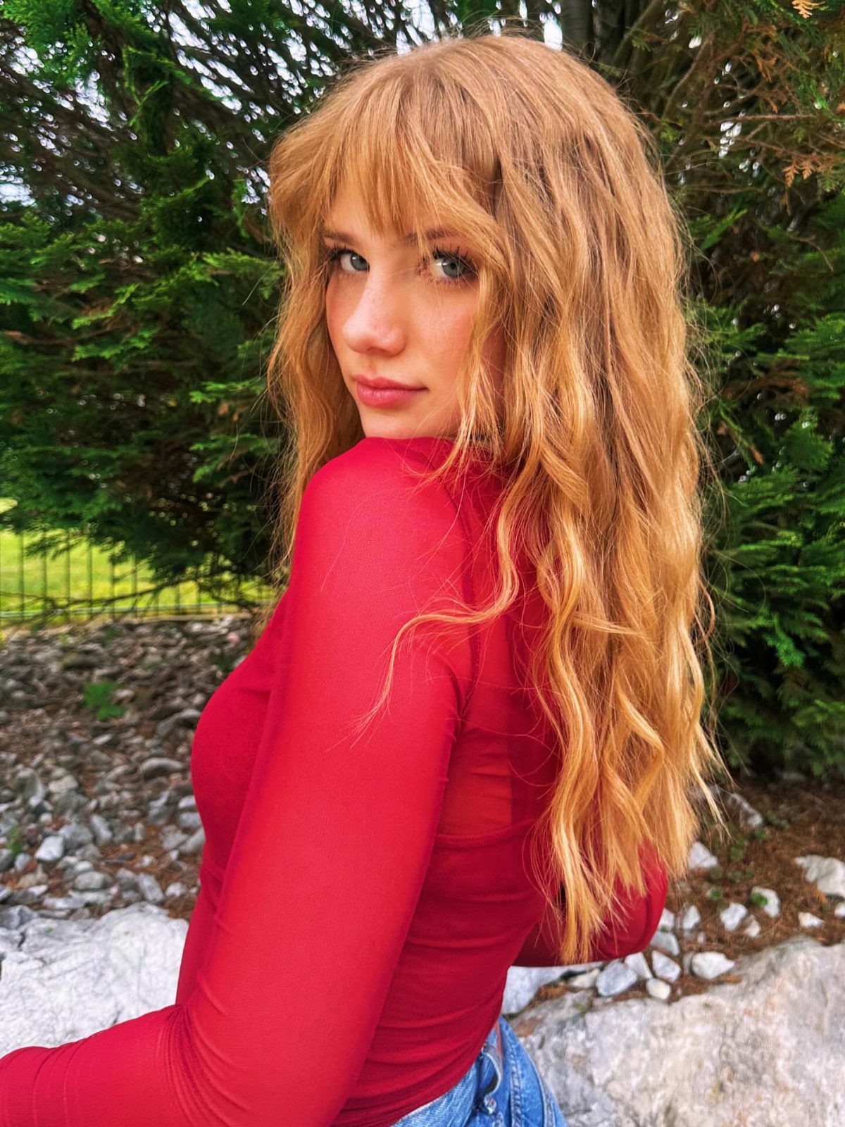 Woman with long, wavy cinnamon strawberry-blonde hair and bangs posing outdoors