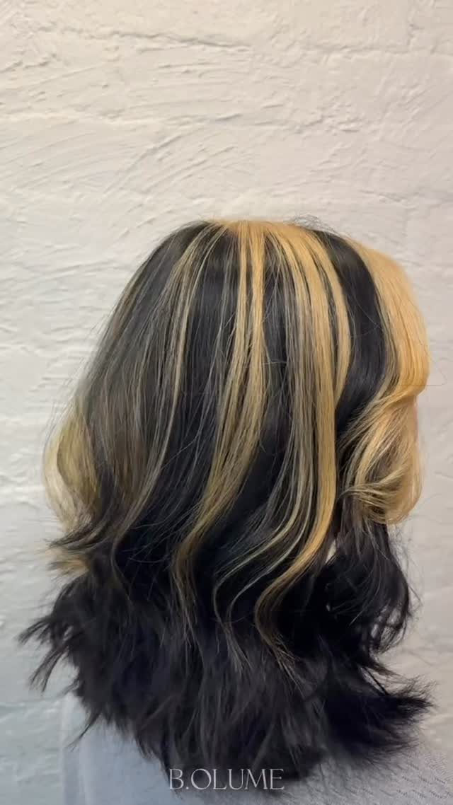 Shoulder-length wavy dark hair with chunky blonde face-framing highlights against a pale wall