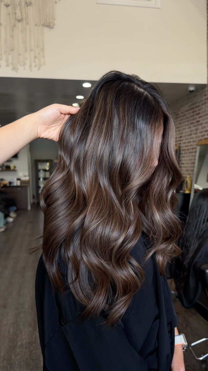 Long dark brown balayage hair falls in glossy waves over a black salon cape