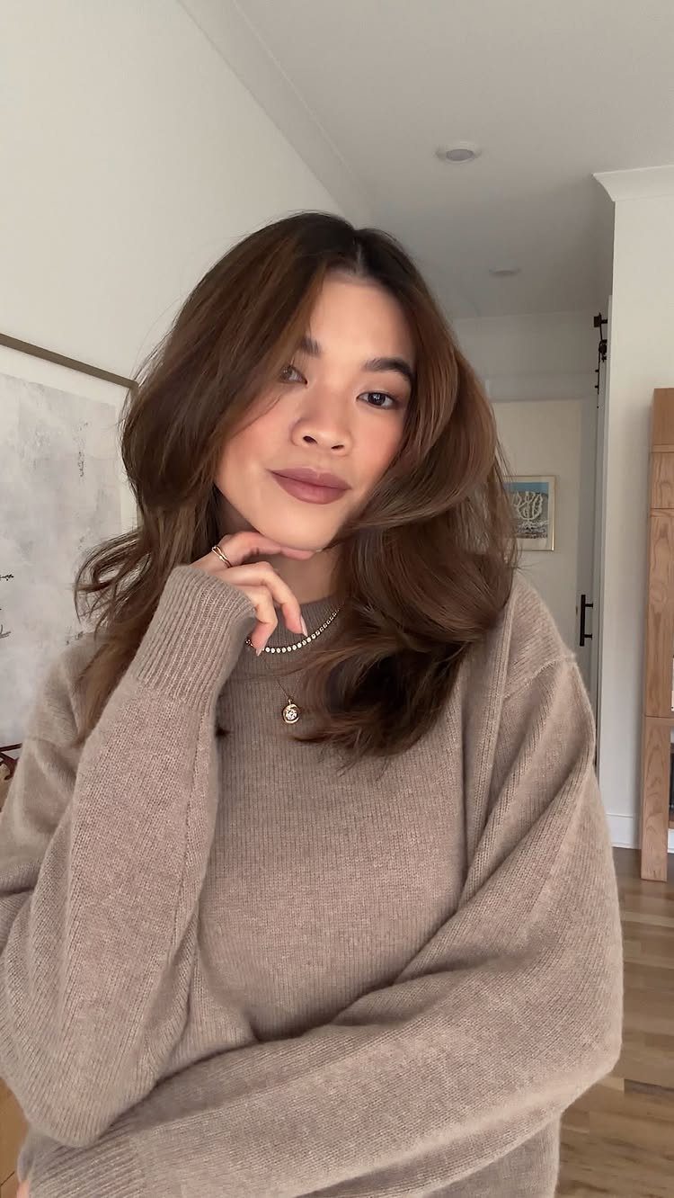 Woman with wavy brunette hair and lighter face-framing strands posing indoors
