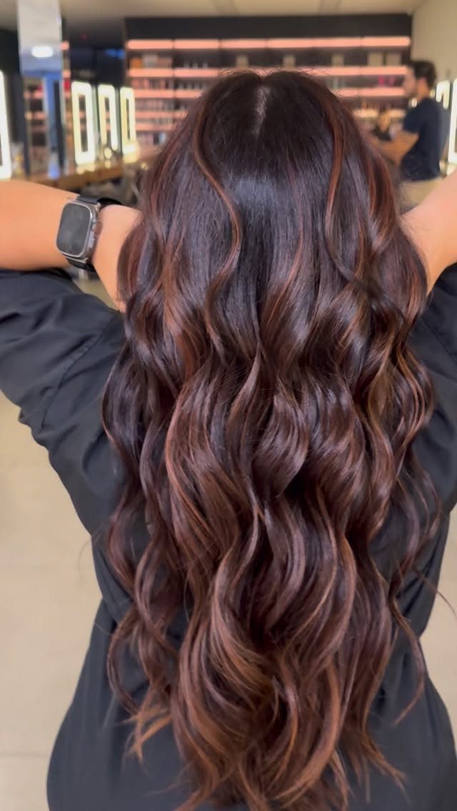 Long dark wavy hair with subtle reddish-brown balayage highlights in a salon