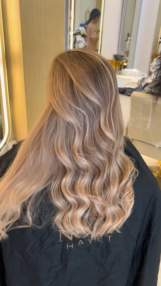 Long blonde balayage waves with darker roots over a black salon cape