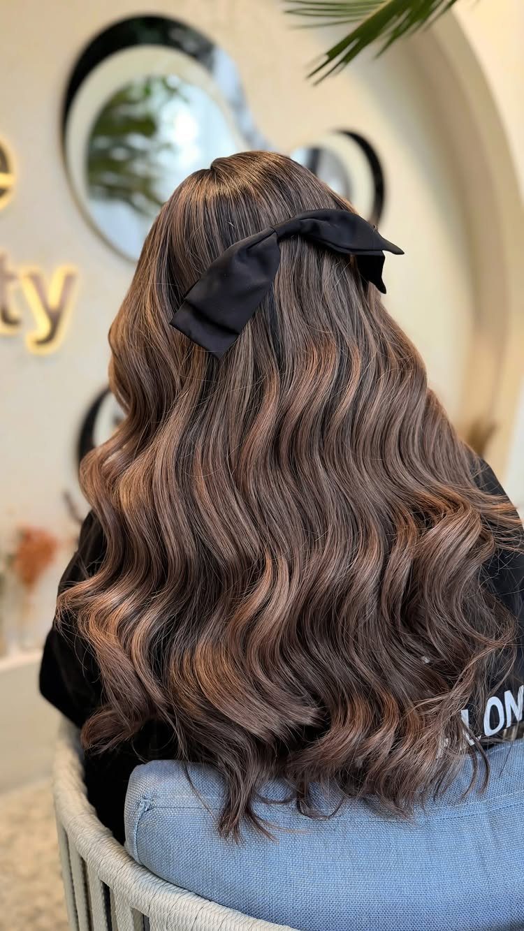 Long brunette balayage waves with lighter brown highlights and a black bow