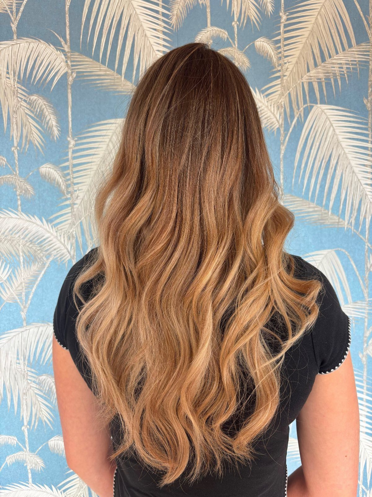 Long brown waves blend into golden blonde balayage against a palm-patterned wall
