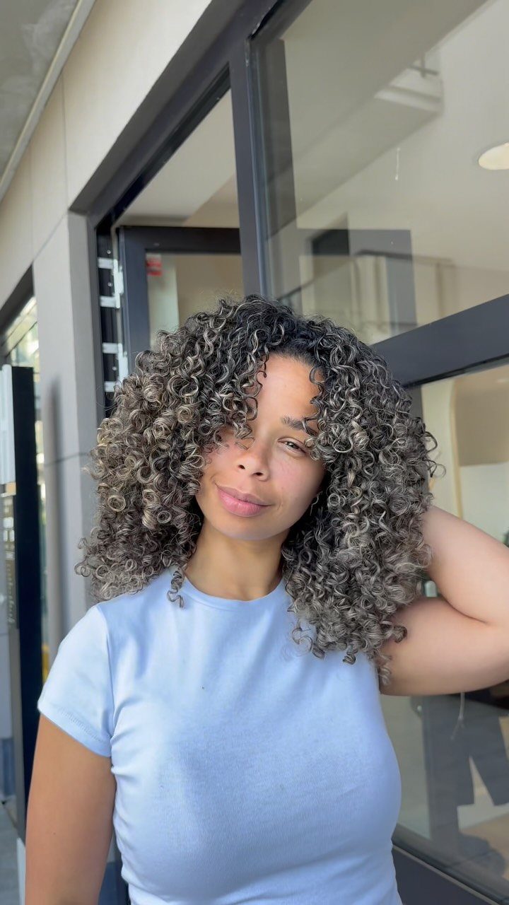 Woman showing ash-blonde highlights throughout her dark curly hair beside a glass doorway