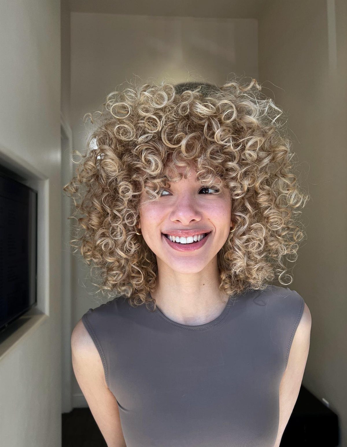 Voluminous blonde curls with darker roots frame a smiling person's face
