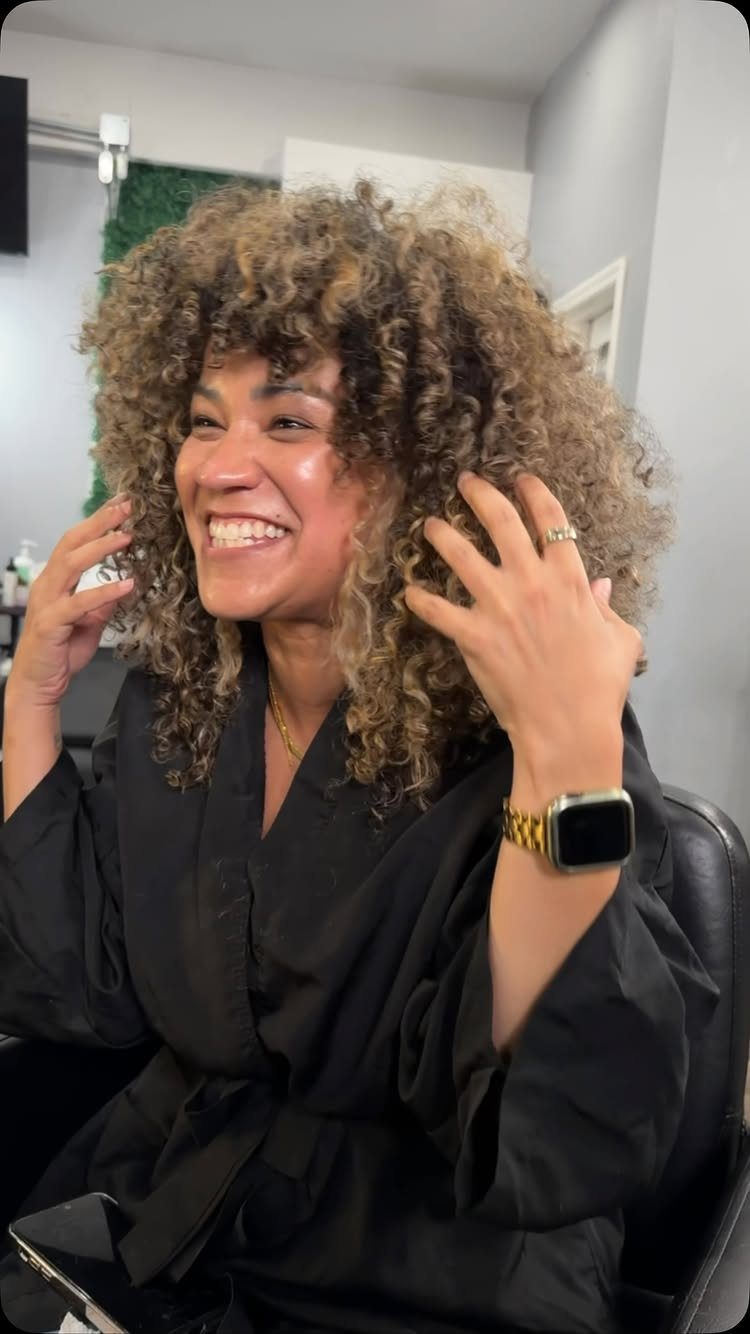 Woman smiling and touching curly brown hair with caramel highlights in a salon