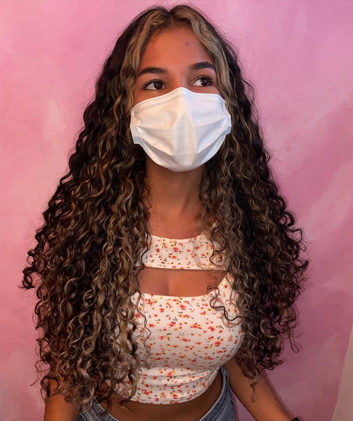 Woman with long dark curls and lighter face-framing pieces against a pink wall