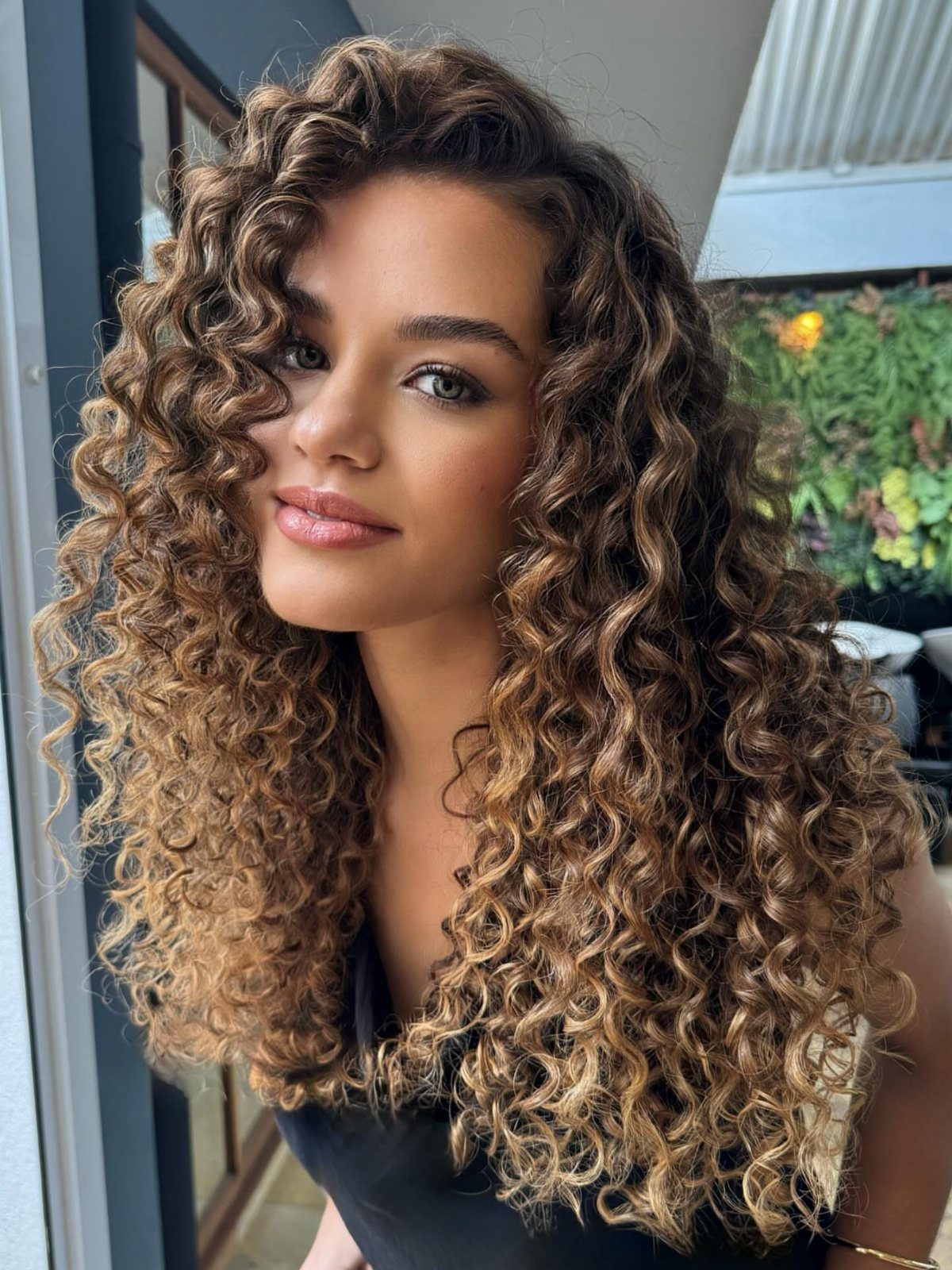 Woman with long brown curls and soft golden ribbon highlights beside a window