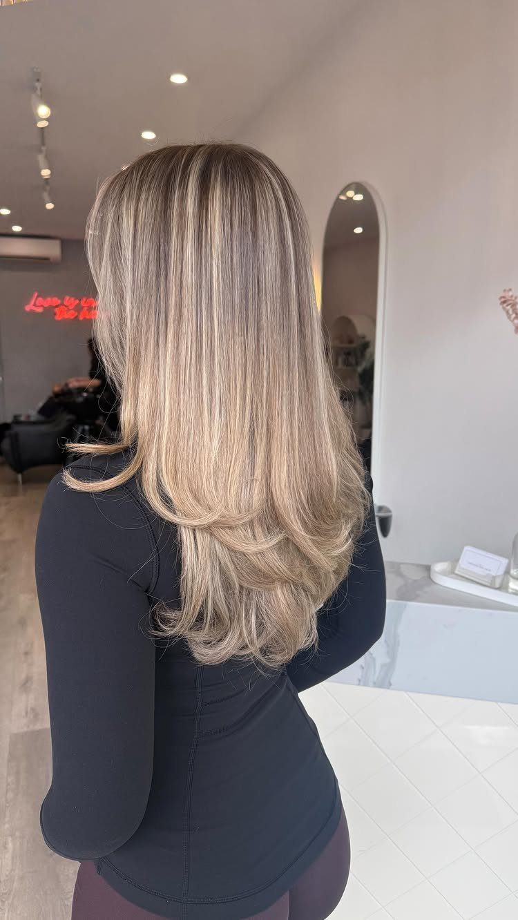 Long layered blonde hair with pale highlights and darker roots in a salon