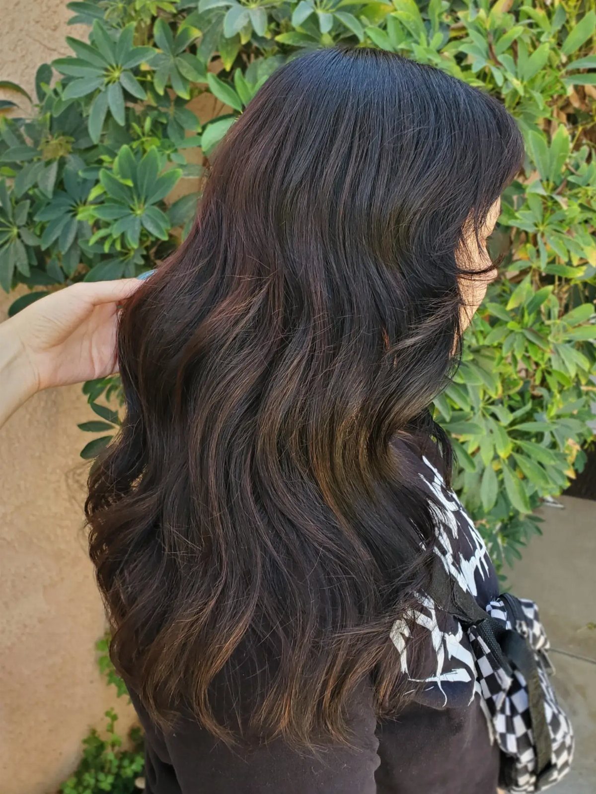 Long near-black wavy hair shows subtle warm brown highlights against green shrubs