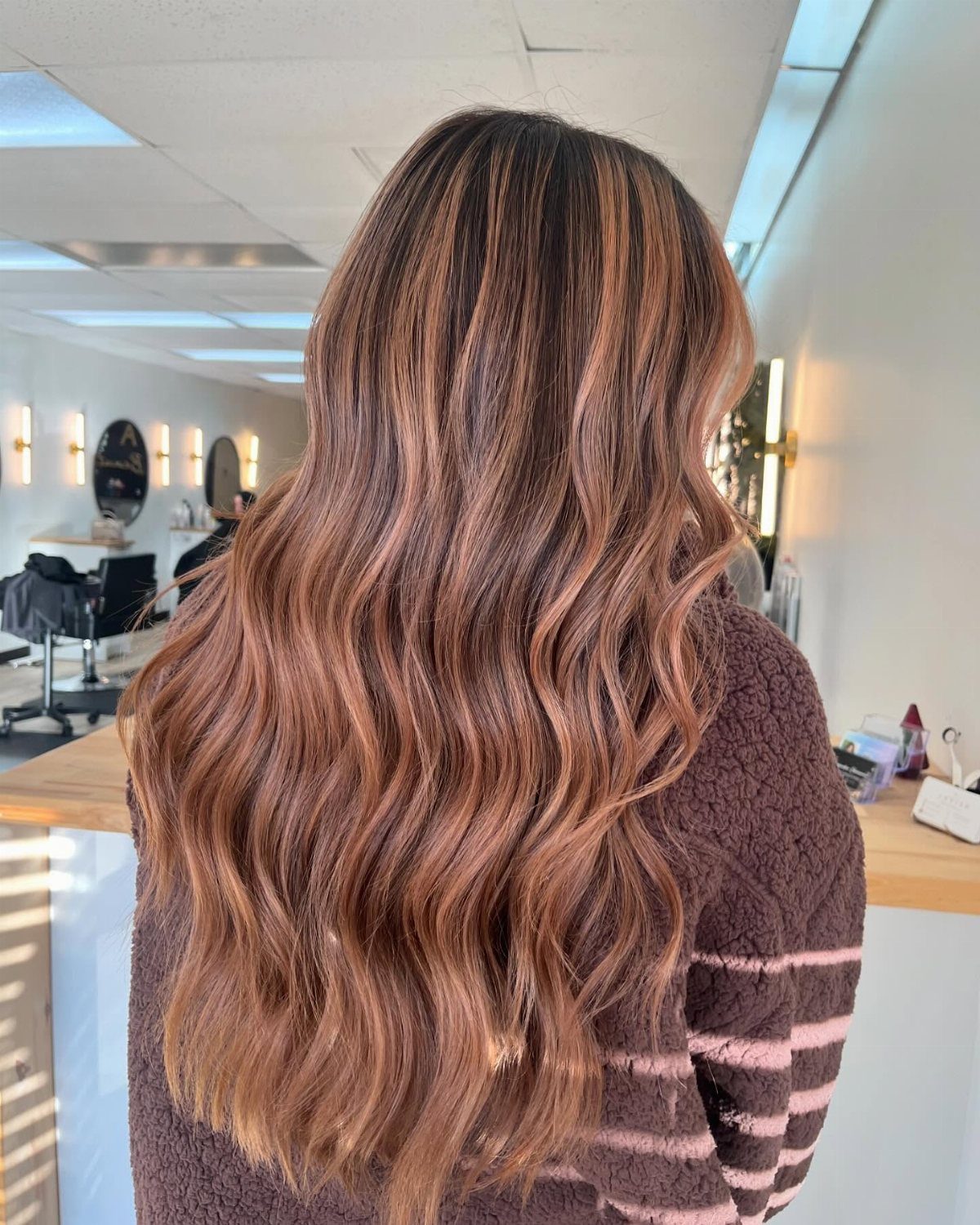 Long wavy brown hair with warm strawberry-blonde balayage highlights in a salon