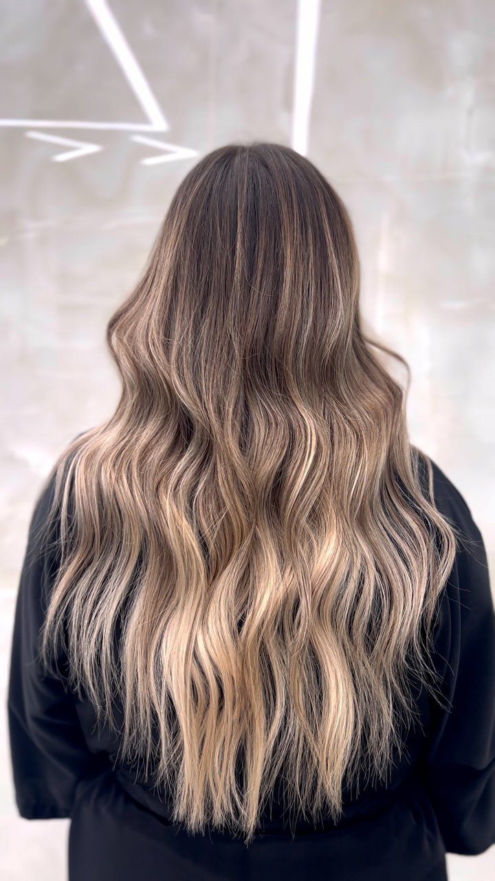 Long wavy brunette hair blends into beige and blonde ends against a pale wall