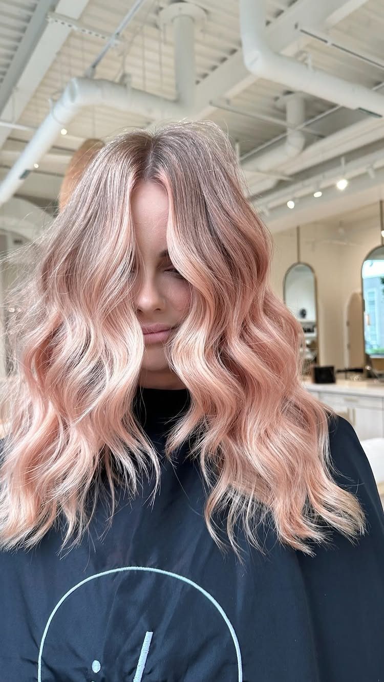 Wavy peach-blonde hair with darker roots frames a person in a salon cape